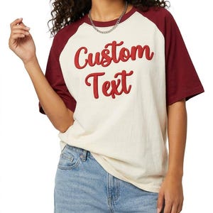 Op de afbeelding: Een crèmekleurig raglan-T-shirt met bordeauxrode mouwen en de woorden Custom Text in verhoogde rode letters op de voorkant. Het T-shirt heeft een ronde hals en korte mouwen, gecombineerd met een lichtblauwe spijkerbroek.