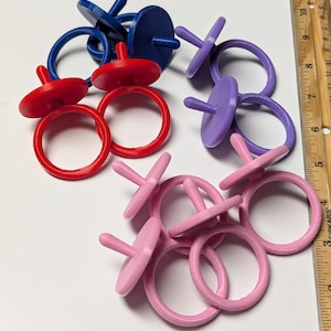 May include: A collection of circular rings with small vertical pegs in red, blue, purple, and pink, arranged next to a wooden ruler showing measurements in inches. The rings are solid-colored and feature a flat disc top with a centered post.