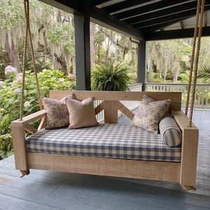 May include: A light-colored wooden porch swing suspended by thick ropes, featuring a blue and white checkered seat cushion, two square floral patterned pillows, one solid beige square pillow, and a blue and white striped cylindrical bolster pillow.