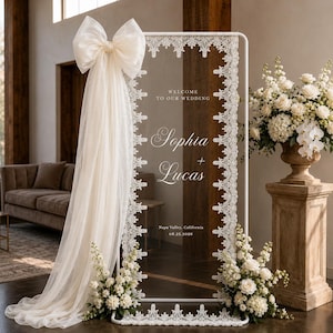 May include: A rectangular white metal frame sign with a clear acrylic panel featuring white lace trim along the edges. The text reads Welcome to our wedding, Sophia + Lucas, Napa Valley, California, 08.25.2026. A large cream tulle bow is attached.