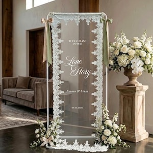 Könnte beinhalten: Ein hohes, rechteckiges Schild aus klarem Acryl mit weißem Metallrahmen und weißer Spitzenborte an den Seiten. Der Text lautet: WELCOME TO OUR Love Story, Emma & Liam, 09.06.2026. Zwei salbeigrüne Bänder sind an den oberen Ecken des Rahmens befestigt.