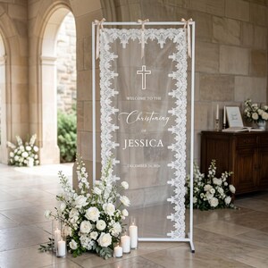 Puede incluir: Un marco de metal blanco rectangular que sostiene un panel de tela blanca transparente con bordes de encaje intrincados. El panel presenta un icono de cruz y texto blanco que dice "WELCOME TO THE Christening OF JESSICA DECEMBER 24, 2026". Tres cintas beige aseguran la tela en la parte superior.
