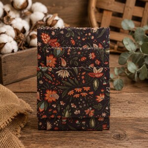 May include: A set of three rectangular beeswax food wraps featuring a dark charcoal background with a repeating floral pattern in shades of orange, cream, and muted green. Each wrap has pinked, zigzag-cut edges and is stacked vertically.