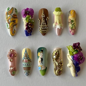 May include: Ten almond-shaped press-on nails featuring intricate designs including a yellow elephant illustration, purple and green floral 3D accents, a gold metallic stupa, a white flower garland, a lotus painting, crystals, and a painted figure.