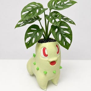 May include: A light green, four-legged creature-shaped planter with red eyes and a red mouth, featuring small green spikes along its back. A green Monstera adansonii plant with characteristic leaf holes grows from the top of the planter.