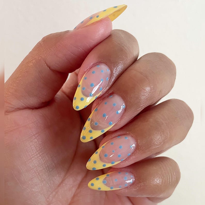 May include: Almond-shaped fingernails featuring a clear base coat with a yellow French tip design. The entire nail surface is decorated with small, light blue polka dots that extend from the yellow tips onto the clear nail bed area.
