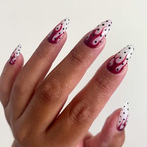 May include: Almond-shaped artificial nails featuring a white base color with a dark red flame pattern and small black polka dots scattered across the surface. The design covers the entire length of each nail, extending from the cuticle to the tip.
