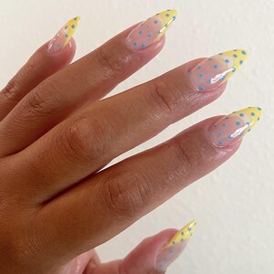 May include: Almond-shaped nails featuring a clear base coat with small, light blue polka dots throughout. The tips of the nails are painted with a solid yellow French manicure style, also decorated with matching light blue polka dots.