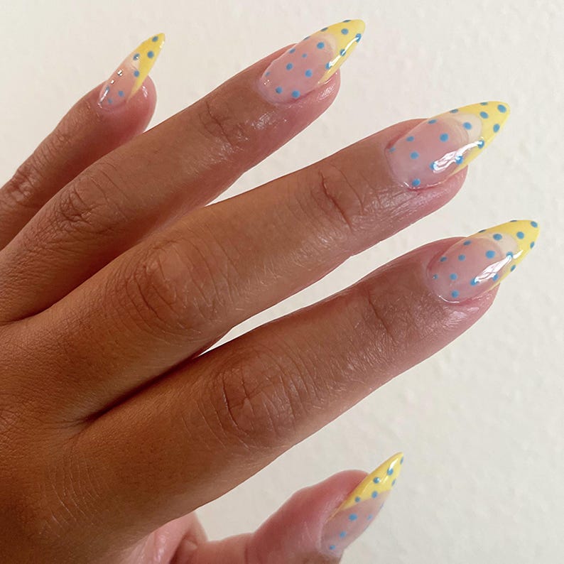 May include: Almond-shaped nails featuring a clear base coat with small, light blue polka dots throughout. The tips of the nails are painted with a solid yellow French manicure style, also decorated with matching light blue polka dots.