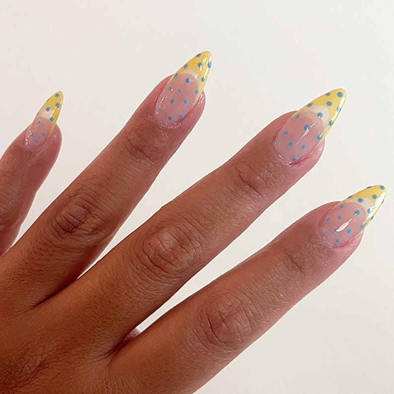 May include: Almond-shaped fingernails featuring a clear base coat with small blue polka dots and a yellow French tip design. The nails have a glossy finish and a smooth, curved shape that extends from the nail bed to a pointed tip.