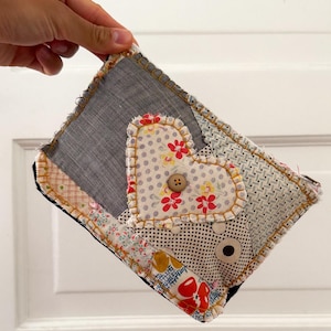 May include: A rectangular fabric pouch featuring a patchwork design with various patterns including gray linen, floral prints, and polka dots. A heart-shaped fabric applique with a central brown button is stitched onto the front with yellow thread.