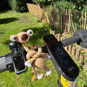 May include: A tan crocheted plush toy resembling a prehistoric squirrel with large white eyes and a dark brown nose, attached to the black handlebar of an electric scooter. The scooter features a digital display screen and a power button.