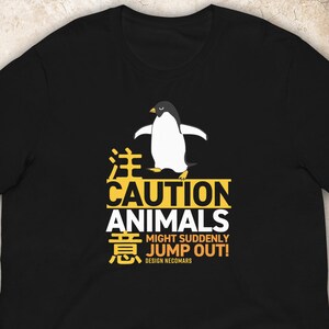 Può includere: Una t-shirt nera a maniche corte con la grafica di un pinguino sopra un testo giallo e bianco che recita: CAUTION ANIMALS MIGHT SUDDENLY JUMP OUT! insieme a caratteri giapponesi e al testo DESIGN NECOMARS stampato in un audace stile a blocchi.