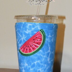 May include: A blue and white fabric cup cozy with a watermelon slice design. The cozy is on a clear plastic cup with a straw.