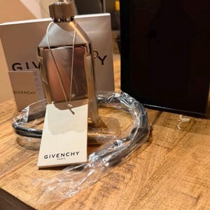 May include: A metallic silver Givenchy bottle with a textured cap and a white tag reading Givenchy Paris. The bottle is partially wrapped in clear plastic, accompanied by a black cord, and positioned in front of a white and black Givenchy box.