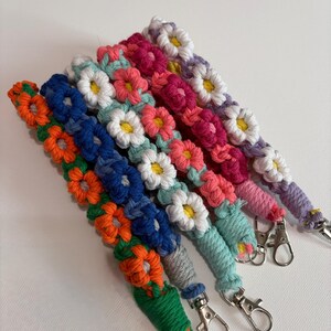 May include: A collection of five macrame lanyards featuring repeating flower patterns in vibrant colors including orange, blue, light blue, pink, and purple, each with a yellow center and a silver metal lobster clasp at the bottom end.