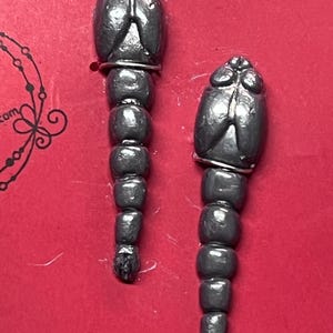 May include: Two dark gray, insect-shaped earrings with a segmented body and a small silver ring at the top. The earrings are displayed on a red background.
