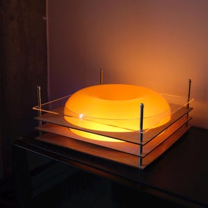 May include: A glowing orange donut-shaped lamp sits centered on a square base composed of three stacked clear acrylic plates. The structure is held together by four vertical silver metal rods with rounded caps at each corner of the square frame.