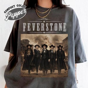 May include: A charcoal gray t-shirt featuring the text COMFORT COLOR Pepper and FEVERSTONE above a graphic of four individuals in western-style black coats and hats standing in front of a burning building. The shirt has short sleeves and a crew neck.