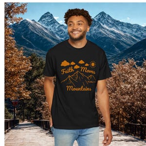 May include: A black short-sleeved t-shirt featuring an orange graphic print of a mountain range, clouds, and a sun. The text "Faith Moves Mountains" is printed in an orange script font, with the small citation "Matthew 17:20" below the peaks.