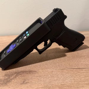 May include: A black plastic remote control holder shaped like a handgun. The holder features a matte finish and is designed to house a television remote with purple and white buttons, including a central purple directional pad and various function keys.