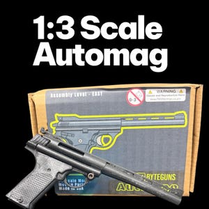 Pode incluir: Um kit de modelo Automag em escala 1:3 na cor preta, com empunhadura e cano texturizados. O produto é exibido em frente à sua embalagem de papelão, que inclui o texto 1:3 Scale Automag, Assembly Level - EASY e uma etiqueta de aviso.