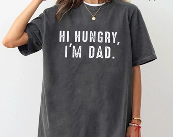 Pode incluir: Uma t-shirt de manga curta em cinzento-carvão com o texto branco "HI HUNGRY, I'M DAD." impresso numa fonte sem serifa desgastada no peito. A peça tem um decote redondo e uma silhueta larga com um acabamento de lavagem vintage.