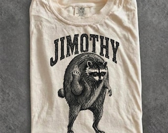 Pode incluir: Uma t-shirt de manga curta cor de creme com uma ilustração gráfica preta de um guaxinim de pé sobre as patas traseiras a levantar o dedo médio. O texto "JIMOTHY" está impresso numa fonte preta, a negrito e arqueada sobre o guaxinim.