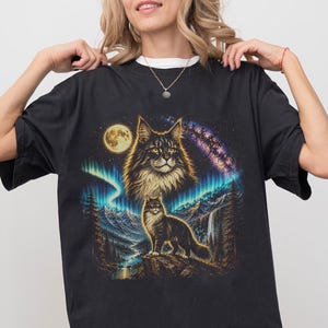 Pode incluir: Uma t-shirt preta com uma estampa gr&aacute;fica de um gato de pelo comprido em p&eacute; num penhasco rochoso, com uma cabe&ccedil;a de gato maior a flutuar por cima. A cena inclui uma lua cheia, uma aurora boreal vibrante, uma gal&aacute;xia e montanhas.
