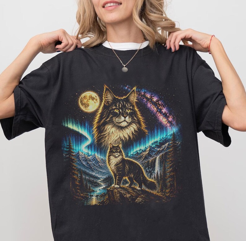 Pode incluir: Uma t-shirt preta com uma estampa gr&aacute;fica de um gato de pelo comprido em p&eacute; num penhasco rochoso, com uma cabe&ccedil;a de gato maior a flutuar por cima. A cena inclui uma lua cheia, uma aurora boreal vibrante, uma gal&aacute;xia e montanhas.