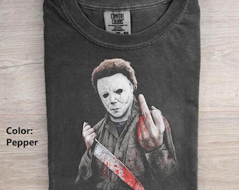 Pode incluir: Uma t-shirt cinzento-carvão com um gráfico de uma figura mascarada com uma máscara branca rachada, segurando uma faca grande com salpicos de sangue vermelho. A figura faz um gesto com a mão com o dedo médio levantado. A etiqueta diz Comfort Colors.