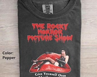 Pode incluir: Uma t-shirt cor de pimenta com o texto vermelho The Rocky Horror Picture Show. Abaixo do texto, há um gráfico de uma pessoa deitada sobre lábios vermelhos grandes. O texto inferior diz Give Yourself Over To Absolute Pleasure.