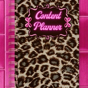 May include: A spiral-bound notebook with a leopard print cover. The cover features the words "Content Planner" in pink script within a decorative frame. The binding is rose gold, and the background is a pink brick pattern.