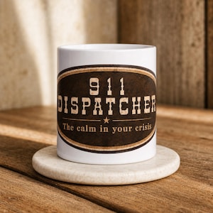 Pode incluir: Uma caneca de cerâmica branca com um gráfico oval castanho-escuro com textura desgastada. O texto diz 911 DISPATCHER e The calm in your crisis, separados por uma pequena estrela. A caneca está sobre um porta-copos redondo de pedra clara.