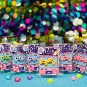 May include: A collection of dangle earrings shaped like video game controllers, each pair displayed on a card featuring a claw machine illustration with the text HoloPop Studio. The controllers are pastel colors including pink, blue, and yellow.