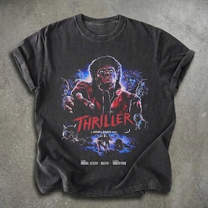 May include: A charcoal gray short-sleeve t-shirt featuring a graphic print of Michael Jackson from the Thriller music video. The design includes the red text Thriller, a John Landis film, and the names Michael Jackson, Ola Ray, and Vincent Price.
