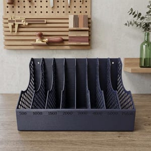 May include: A dark blue desktop organizer with seven vertical slots and a mesh side design. The front base features printed numerical labels 500, 1000, 1500, 2000, 3000, 4000, 5000, and the word Polish, indicating a storage system for abrasive sanding sheets.