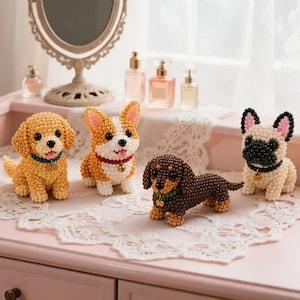 以下が含まれることがあります： 小さなビーズで作られた4匹の犬の置物。左から順に、青い首輪をしたゴールデンブラウンの子犬、赤い首輪をした白と茶色のコーギー、緑の首輪をしたダークブラウンのダックスフント、そしてクリーム色のフレンチブルドッグが並んでいます。