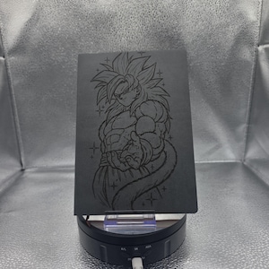 Peut inclure: Un carnet rectangulaire gris foncé présentant une illustration gravée au laser d'un personnage d'anime aux cheveux en pointe dans une posture de combat. Le personnage est entouré de petites étoiles. Le carnet est posé sur un support rotatif noir.