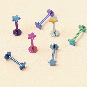 May include: A collection of six labret body jewellery pieces featuring star-shaped tops. Each piece is crafted from iridescent metal, displaying a spectrum of colours including blue, purple, green, and gold, with a flat circular base at one end.