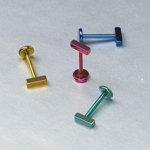 May include: Four metal labret studs featuring a flat circular base and a horizontal rectangular bar top. The studs are finished in metallic gold, blue, magenta, and teal colours, each with a smooth, polished surface and a straight post.