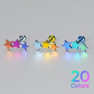 May include: Three metal stud earrings featuring a horizontal design of two connected stars of varying sizes. Each earring displays a vibrant, multi-coloured gradient finish, transitioning through shades of blue, purple, yellow, and green. Text reads 20 Colours.