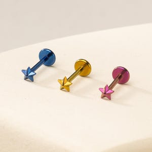 May include: Three labret body jewellery pieces featuring star-shaped ends. Each piece has a flat circular back and a straight post. The first is metallic blue, the second is metallic gold, and the third is a metallic pink and purple gradient finish.