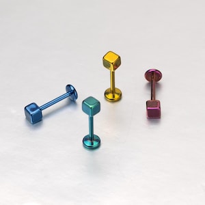 Può includere: Quattro piercing labret con estremità a forma di cubo e finiture metalliche. Il set include un pezzo blu, uno turchese, uno dorato e uno viola. Ogni elemento è composto da una base circolare piatta, un perno dritto e una parte superiore quadrata.