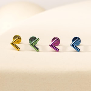 May include: Four stud earrings featuring a chevron or arrow-shaped design. Each earring has a flat circular back and a metallic finish in distinct colors: gold, green, purple, and blue. The studs are arranged in a horizontal row.