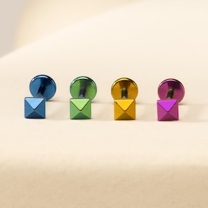 May include: Four labret studs featuring square pyramid-shaped heads in metallic blue, green, gold, and purple. Each stud has a flat circular back disc and a straight post, showcasing a polished finish that reflects light across the four distinct colours.