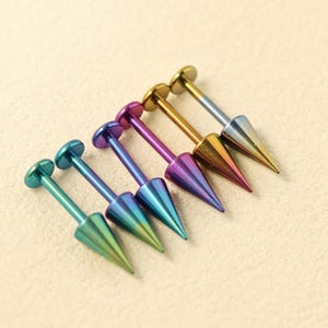 May include: A set of six labret body jewelry pieces featuring flat circular backings and pointed cone-shaped fronts. The jewelry displays a gradient of metallic colors including teal, blue, purple, gold, and silver, arranged in a row.