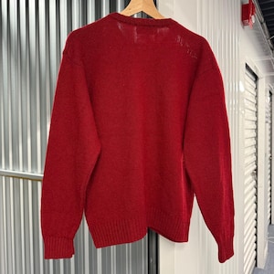 May include: A solid red long-sleeve knit sweater featuring a crew neckline and ribbed cuffs at the wrists and hem. The garment is displayed on a wooden hanger, showcasing a consistent deep red color throughout the entire knitted fabric texture.