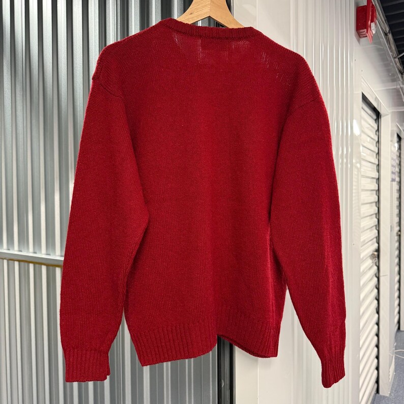 May include: A solid red long-sleeve knit sweater featuring a crew neckline and ribbed cuffs at the wrists and hem. The garment is displayed on a wooden hanger, showcasing a consistent deep red color throughout the entire knitted fabric texture.