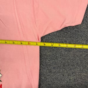 May include: A light pink t-shirt featuring a partial graphic of a flower with white petals and a yellow center. A yellow measuring tape is positioned horizontally across the chest, showing a width measurement of approximately 11 inches.